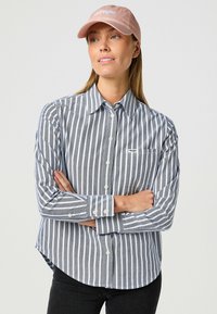 Camisa de botones a rayas grises y blancas con bolsillo y dobladillo curvado. Combinada con una gorra rosa claro que presenta texto bordado.