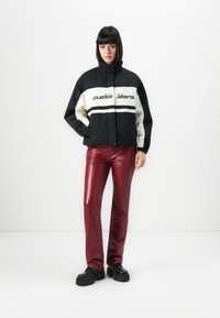 Fekete, egyenes hajú nő fekete-fehér Guess Jeans dzsekit, fényes piros bőr nadrágot és fekete vastag talpú cipőt visel, egy fehér fal előtt áll.