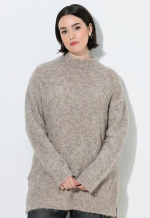 Ung kvinde med kort mørkt hår iført en beige blød sweater, stående mod en hvid baggrund med hænderne blidt foldet.