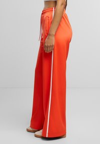 Orangene weit geschnittene Hose mit einem Kordelzug-Bund und weißen Seitenstreifen. Hergestellt aus glattem, leichtem Material. Flache Taschen an den Hüften.