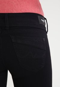 Acercamiento de una persona que lleva jeans negros con detalles de costura en el bolsillo trasero y etiqueta de la marca Pepe Jeans en la cinturilla.