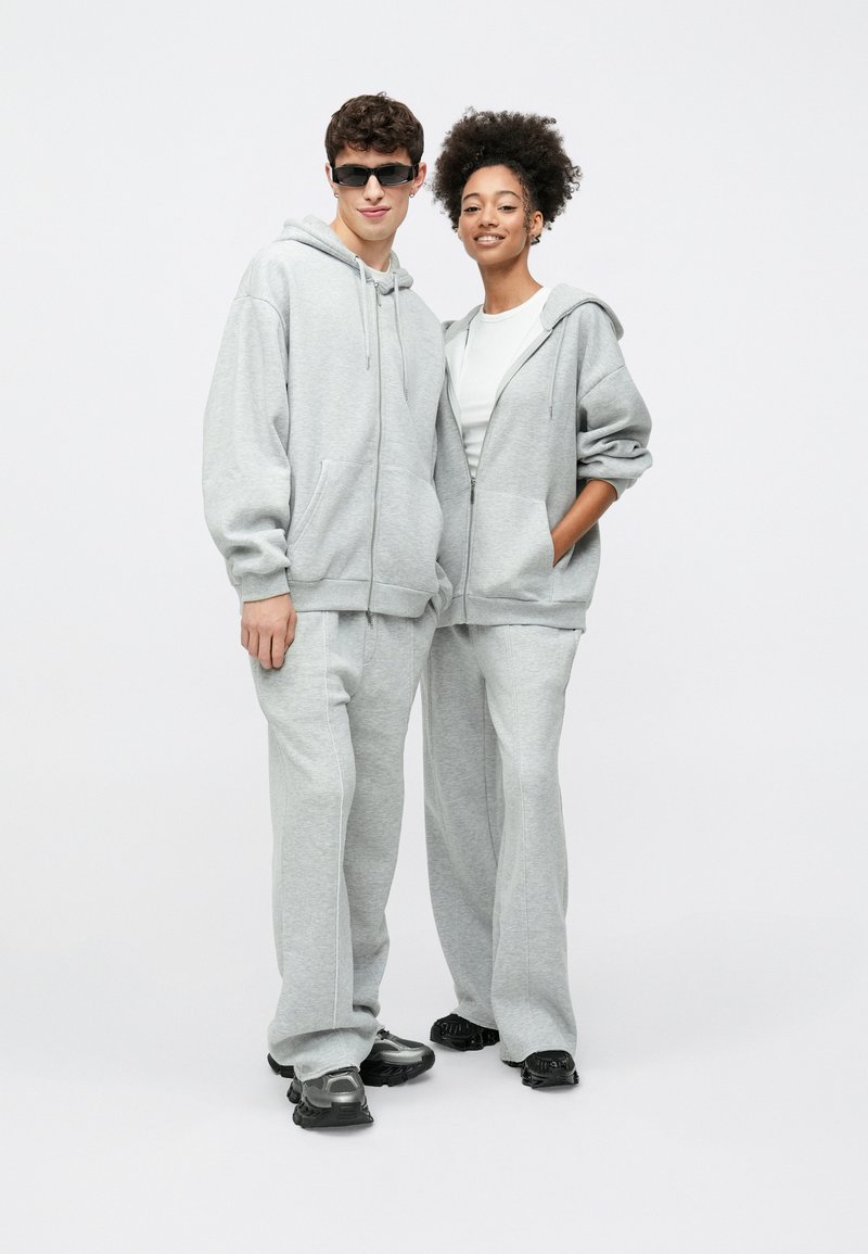 Pilki gobtavai megztiniai su gobtuvu ir treningai iš minkštos medžiagos, turintys kišenes kengūros stiliaus, laisvo kirpimo ir kontrastingas baltas siūles. Avimi juodi sportbačiai.