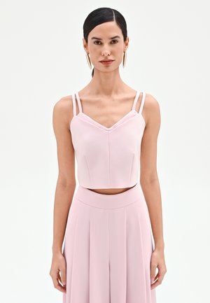 Donna con capelli scuri raccolti indossa grandi orecchini a cerchio dorati e un top corto rosa chiaro con doppia spallina abbinato a pantaloni a vita alta coordinati.