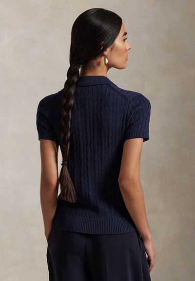 Kobieta z długimi splecionymi warkoczami, ubrana w granatowy, krótki sweter z warkoczowym splotem, widoczna od tyłu na jednolitym tle.