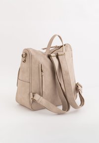 Mochila de cuero beige con correas ajustables, asa superior, herrajes dorados y dos bolsillos laterales; textura suave y diseño estructurado.