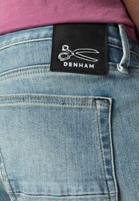 Vaqueros Denham con un lavado de denim en azul claro, con una etiqueta negra que lleva el logo de tijeras blancas y detalles de costura en los bolsillos.