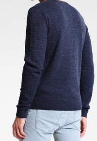 Homme portant un pull texturé bleu marine et un jean bleu clair, vu de dos sur un fond uni.