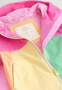 Otroška jakna v pastelnih barvah z rumenim zadrgo, rožnato kapuco in notranjo etiketo z napisom »Little Bird« ter prostorom za vpis imena lastnika.