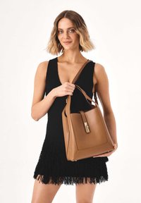 Handtas van lichtbruin leer met een gestructureerde rechthoekige vorm, afneembare band en metalen sluiting, gedragen door een model met een zwarte jurk met franjes.