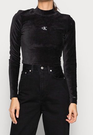 Person iført sort fløjls langærmet crop top med hvid "cK" logo og højtsiddende sorte jeans, stående mod en ensfarvet baggrund.