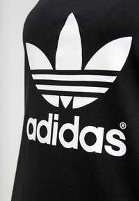 Svart bomullströja med en stor vit Adidas-logga med tre ränder och trefoil-design. Texten "adidas" i fetstil vita bokstäver.