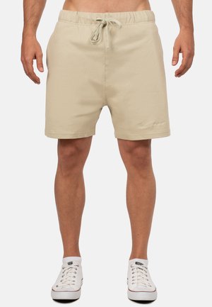 Mężczyzna w beżowych szortach ze sznurkiem i białych niskich tenisówkach, stojący na jednolitym jasnym tle.