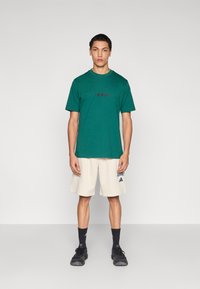 Zelené bavlnené tričko s okrúhlym výstrihom a čiernym logom „adidas“, v kombinácii s béžovými šortkami s logom. Nosené s čiernymi teniskami a ponožkami.