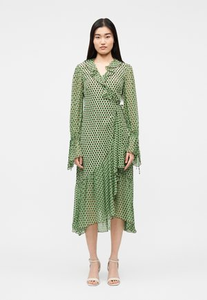 Woman wearing a green long-sleeve midi dress with brown polka dots, ruffled collar, and asymmetrical hem, paired with white ankle-strap heels.