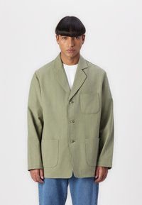 Olive green jacket with a notched collar, three buttons, and two front pockets. Smooth fabric with slight texture, paired with blue denim jeans.