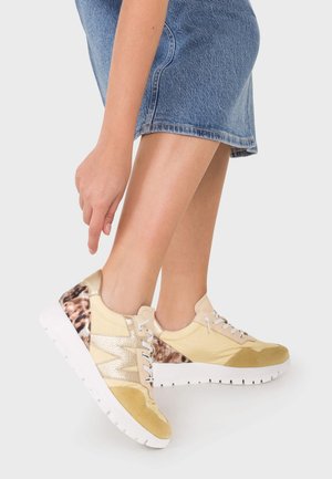 Persona che indossa sneakers gialle e dorate con suola bianca e dettagli con stampa leopardata, abbinate a una gonna di denim blu.