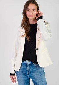 Blazer crème avec un bouton noir, coupe ajustée, revers en pointe, porté sur une chemise noire, associé à un jean bleu. Tissu lisse, design minimal.
