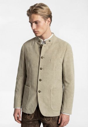 Junger Mann trägt eine beige Cordjacke mit Stehkragen und Knöpfen sowie braune Lederhose mit Stickereien.