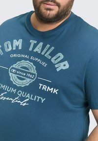 Cyan katoenen t-shirt met ronde hals, voorzien van witte bedrukte branding en grafisch ontwerp. Korte mouwen en zachte textuur zijn zichtbaar.