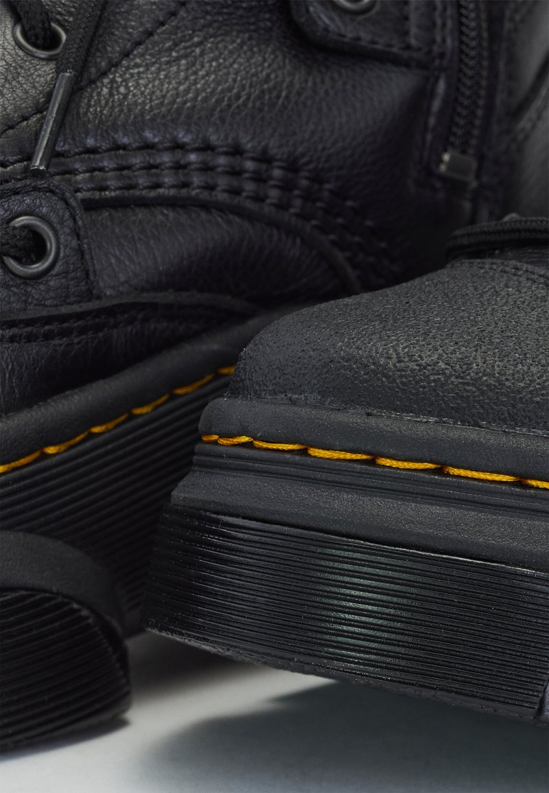Bottes en cuir noir avec surface texturée, bout rond, surpiqûres jaunes et semelles en caoutchouc. Présentent des œillets ronds pour les lacets.