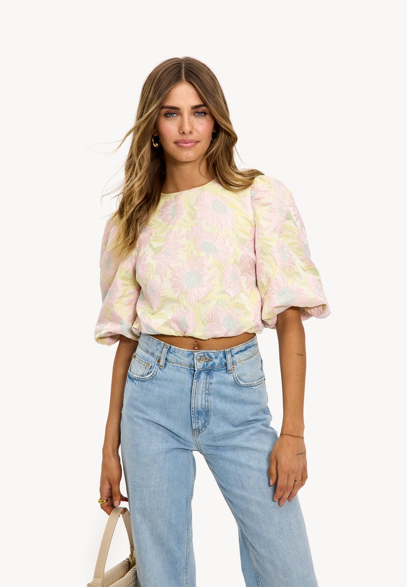 Femme portant un crop top à manches bouffantes à fleurs pastel et un jean taille haute bleu clair, tenant un sac à main beige à ses côtés.