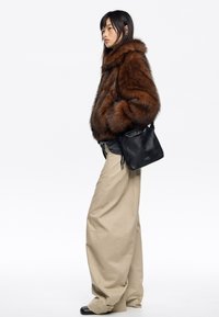 Žena v hnedej umelohovratnovej bunde, béžových širokých nohaviciach, čiernych topánkach a čiernej kabelke, stojaca bokom na bielom pozadí.