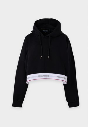 Γυναίκα που φοράει μαύρο cropped hoodie και τζιν με φαρδιά πόδια με ασπρισμένες κηλίδες και μικρά σκισίματα, στέκεται μπροστά σε ένα απλό φωτεινό φόντο.
