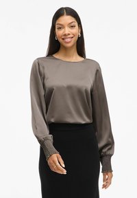 Brun satin bluse med lange ærmer, sammensnørede manchetter, rund halsudskæring og en glat tekstur, parret med en tætsiddende sort nederdel.