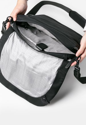 Handen openen een zwarte schoudertas en tonen een ruime, grijs patroon binnenkant met meerdere vakken en een ritssluiting.