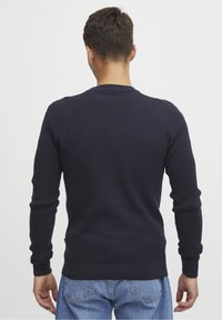 Homme portant un pull à manches longues texturé bleu marine et un jean bleu, debout dos à l'appareil photo devant un fond uni.