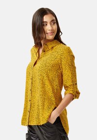 Yellow button-up shirt with black polka dots, long sleeves rolled up, collar design, and side slits for a loose fit.