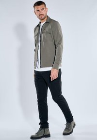 Leichte olivgrüne Jacke mit zwei Brusttaschen, kombiniert mit schwarzen Cargo-Hosen und grauen Stiefeln, vor einem neutralen Hintergrund.