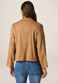 Bruine cropped jas met lange mouwen, knoopaccenten op de mouwen en een relaxte pasvorm. Draagt over een blauwe denim jeans. Glad stofoppervlak.