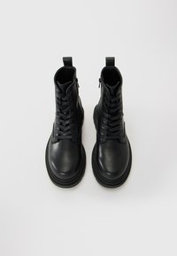 Par de botas de combate de cuero negro con cordones, cremalleras laterales y tiradores, fotografiadas desde arriba sobre un fondo blanco.