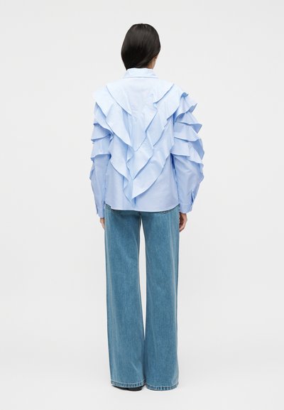 Γυναίκα που στέκεται γυρισμένη με την πλάτη φορώντας ανοιχτό μπλε μπλούζα με βολάν και φαρδιά μπλε τζιν σε απλό ανοιχτό φόντο.