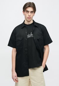 Νέος άνδρας με σκουλαρίκια, που φοράει μαύρο πουκάμισο με κοντά μανίκια και κουμπιά πάνω από μαύρο μπλουζάκι με στάμπα και μπεζ σορτς, σε λευκό φόντο.