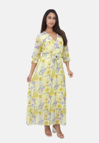 Maxikjole i gul med blomster mønster, der har gule og lilla blomster. V-hals, flæseærmer og stofbælte i taljen.