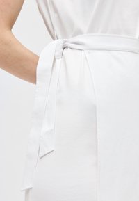 Gros plan sur une robe blanche avec une ceinture en tissu nouée autour de la taille et le bras d'une personne reposant à côté.
