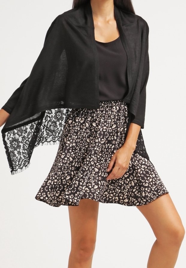 Femme portant un haut noir, un châle en dentelle noire et une jupe courte noire à motif floral blanc, debout avec une main sur la hanche.