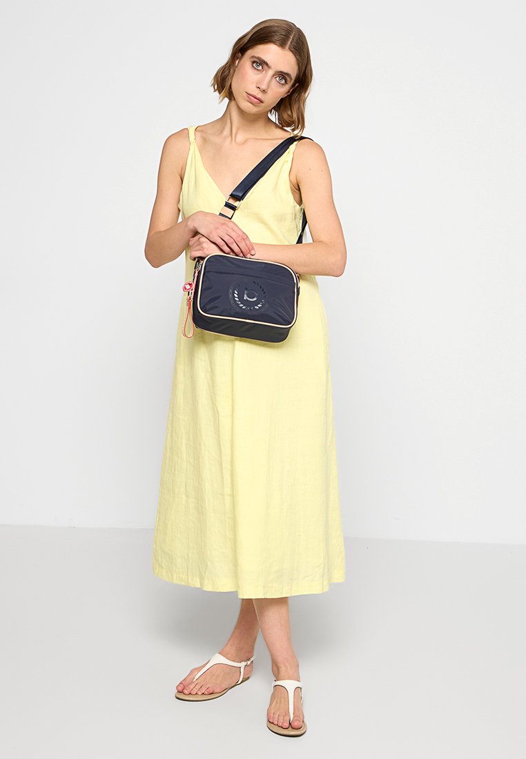 Sac rectangulaire marine avec finition crème et design de logo, doté d'une bandoulière et d'une fermeture zippée, présenté avec une robe jaune.
