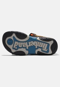 Suela de goma negra y azul con patrón texturizado, con acentos blancos y la marca "Timberland" en letras grandes. Detalle de correa ajustable.