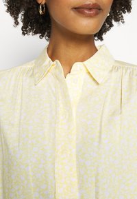 Lichtgele blouse met een kraag, voorzien van een wit bloemenpatroon. Getextureerde stof met plooien langs de schouders en een knoopsluiting aan de voorkant.