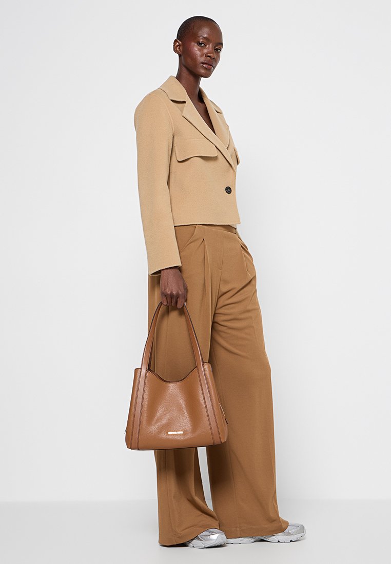 Beige cropped jas met twee voorzakken, gecombineerd met tan wijde broek. Model houdt een bruine leren draagtas met een gladde textuur vast.