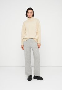 Μπεζ πλεκτό πουλόβερ με ψηλό λαιμό, συνδυασμένο με γκρι πλεκτό παντελόνι με φαρδιά μπατζάκια. Το μοντέλο φοράει μαύρα παπούτσια με χοντρή σόλα. Απλό λευκό φόντο.