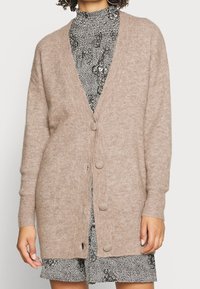 Beige stickad cardigan med V-ringning, långa ärmar och sex knappar. Mjuk textur med sidfickor. Buren över en svart och vit mönstrad klänning.