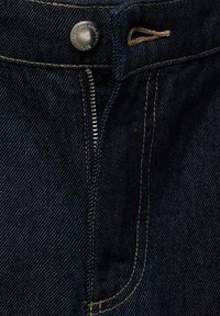 Des jeans en denim foncé avec une fermeture éclair à l'avant, un bouton métallique rond et des coutures beiges contrastantes le long des bords et des poches.