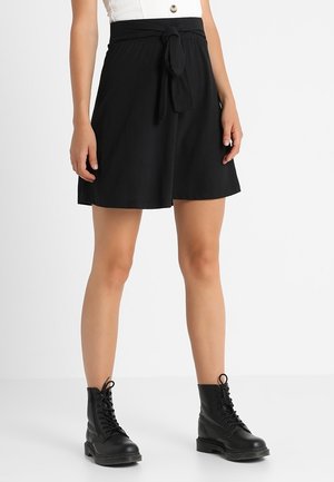 Kobieta ubrana w czarne szorty z wysokim stanem i pasek wiązany, białą bluzkę oraz czarne, sznurowane botki, stojąca na jednolitym białym tle.