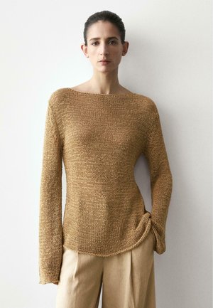Donna che indossa un maglione lavorato a maglia marrone chiaro e ampio e pantaloni beige a gamba larga, in piedi contro una parete liscia e chiara.