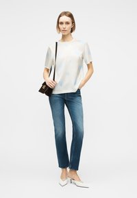 Γυναίκα που φορά λευκό τοπ με κοντά μανίκια, μπλε τζιν, λευκές μυτερές γόβες και κρατά μαύρη τσάντα ώμου, με το ένα χέρι στην τσέπη.