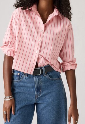 Frau trägt ein rosa langärmeliges Hemd mit roten Streifen, hochgekrempelten Ärmeln, in eine hoch taillierte blaue Jeans mit schwarzem Gürtel gesteckt.